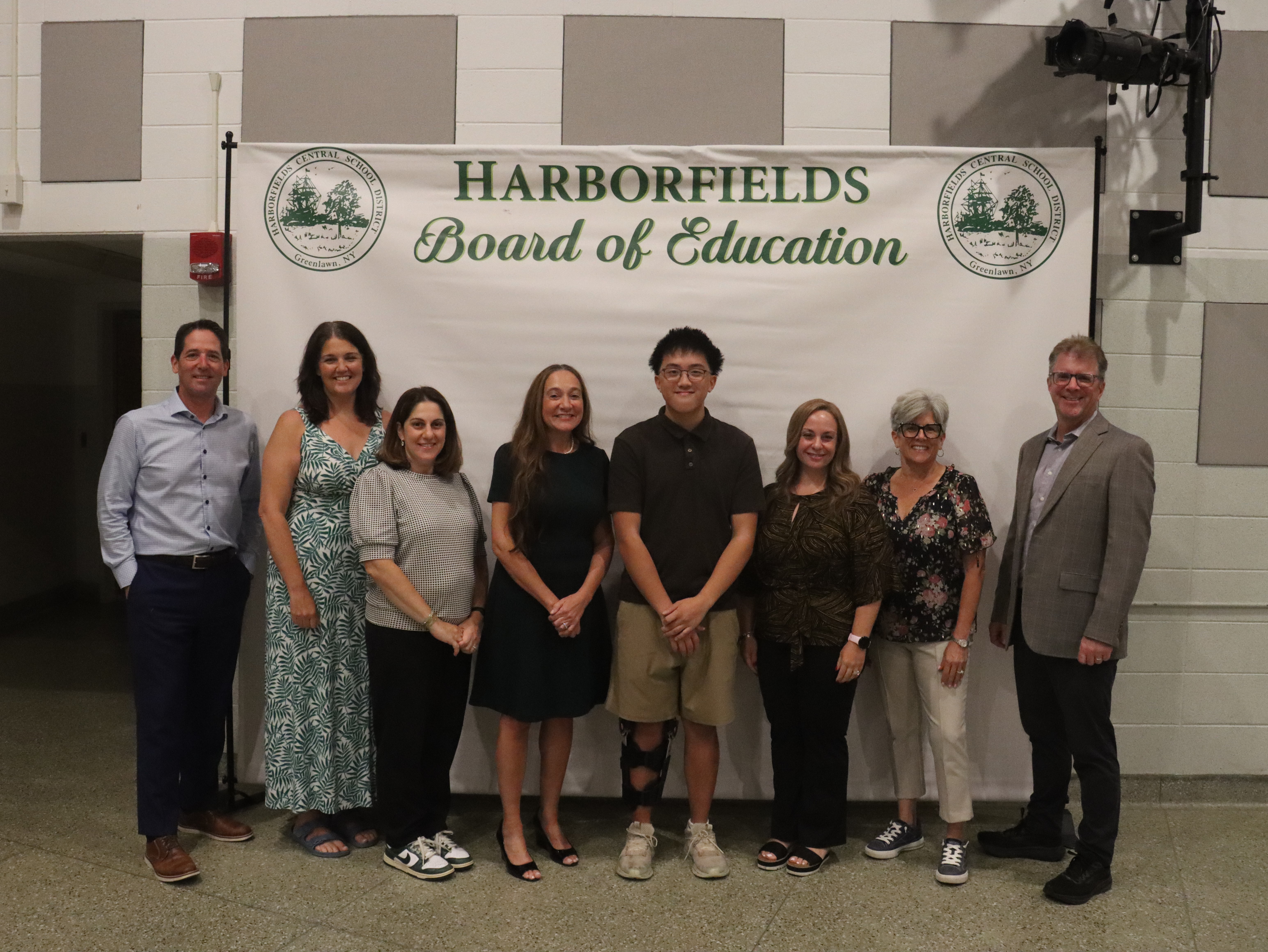 A group of eight people stand in front of a banner that reads "HARBORFIELDS Board of Education".